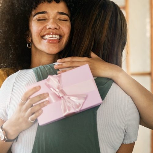 A woman smiles excitedly as she hugs another woman at home, while holding a gift wrapped with a pink ribbon.