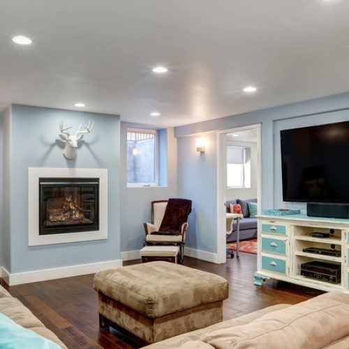 A modern finished basement features wooden flooring, a brown sectional, a mounted television, and a painted dresser.