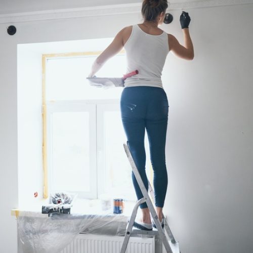 A woman stands on a ladder and uses a small brush to paint a wall in her home. She holds a paint tray with her other arm.