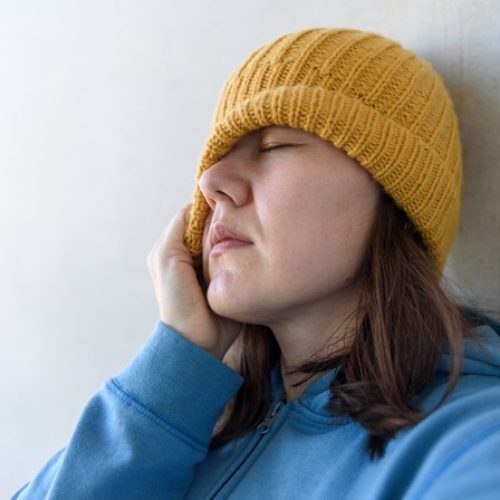 An anxious woman in a blue pullover tugs a yellow beanie over her right eye and leans against a white wall.