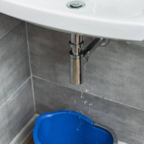 A white floating sink has exposed silver pipes underneath it. Water is leaking from the pipes into a blue bucket.