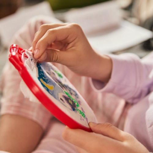 A close-up of a woman stitching a colorful pattern into white fabric in a red embroidery hoop while sitting in pink pajamas.