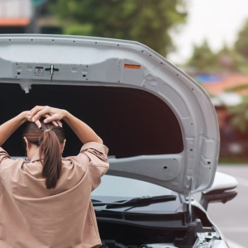 The back of a woman looking under the hood of her car. She has her hands on her head in frustration.