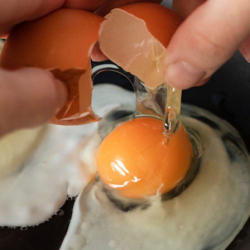 A close-up view shows a pair of hands opening an egg with a brown shell over a skillet. Another egg cooks in the pan.