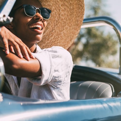 Four smiling women walking arm-in-arm outdoors. Three wear glasses, and two are in wide-brimmed straw hats.