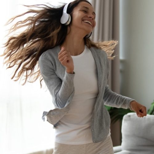 A young woman with long brunette hair wears a pair of white headphones as she dances while smiling widely.