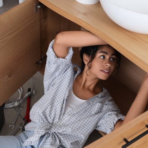 A woman lays under a sink in a laundry room and is repairing the plumbing with tools and supplies next to her.