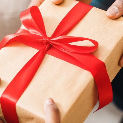 One pair of hands giving a wrapped present to another pair of hands. The gift is wrapped in brown paper and red ribbon.