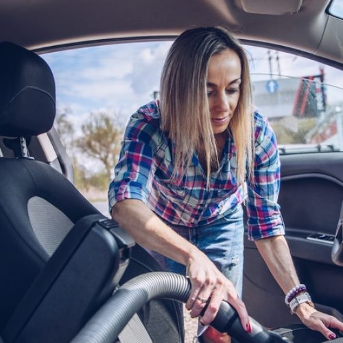 A blonde woman is vacuuming the front seat of her car at a car wash while the sun shines on a bright day.