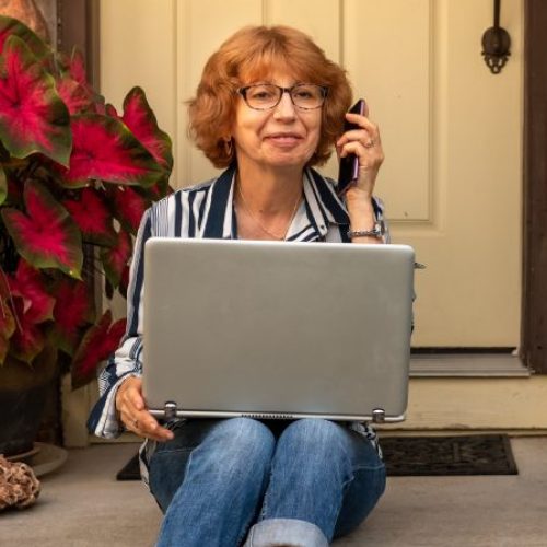A woman sits on her front porch next to a large plant. She holds a laptop in her lap and has a phone next to her ear.