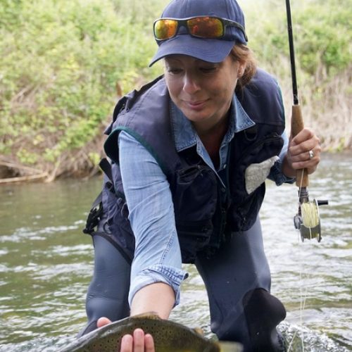 A woman in blue fishing gear holds a rod in one hand and a fish in the other, bending slightly to pull it from the water.