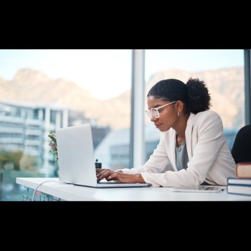 A focused young woman wearing glasses and a blazer typing on a laptop while working at a desk near a large picture window.