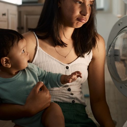 A young woman sits on the floor in front of a washing machine while holding a toddler and looking frustrated.