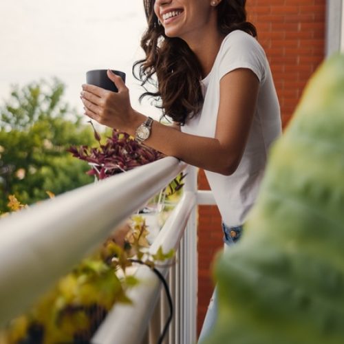 A woman stands on her balcony holding a coffee mug and smiles as she looks out over trees and greenery in the distance.