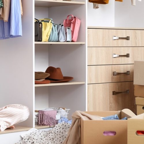 A young woman sitting in a large closet wearing a hat, with boxes of clothing stacked up and clothing scattered around.