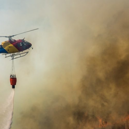 A rescue helicopter flying over a wildfire zone with a water tank dangling from the chopper. There are flames and smoke.