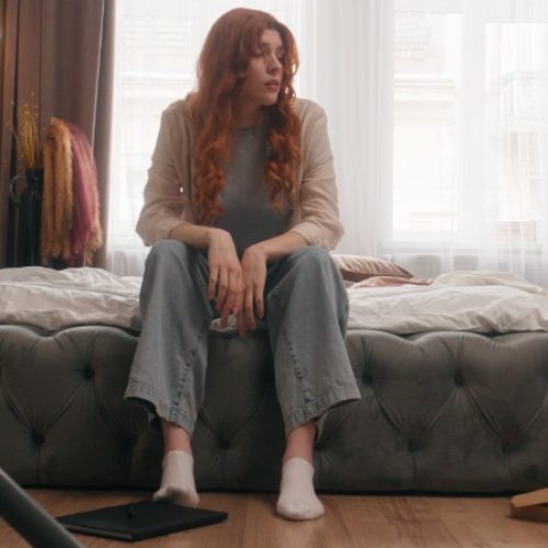 A woman sits on the edge of an unmade bed in a cluttered bedroom, surrounded by household items and a vacuum cleaner.