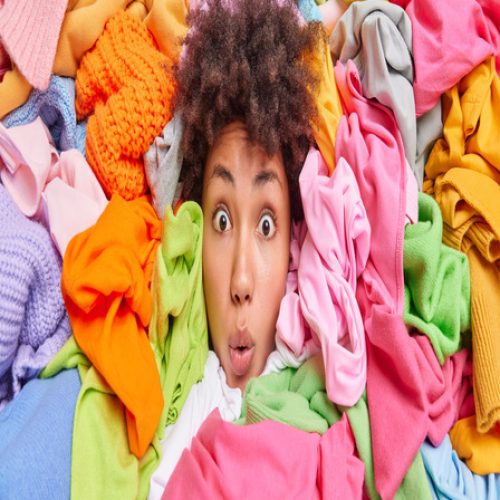 A surprised woman’s face peeks out from a vibrant pile of colorful clothes in pink, green, yellow, and blue hues.