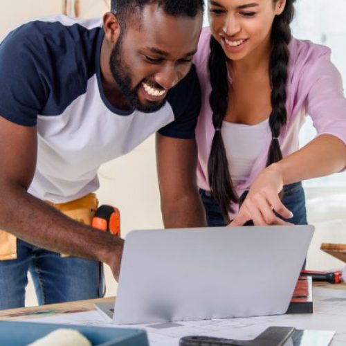 A man and woman look over a laptop set on a full work table. They grin, excited to work on their home expansion.