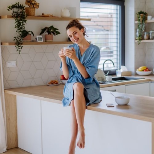 A woman in a robe sitting on a kitchen counter holding a mug, smiling in a bright kitchen with plants and shelves.