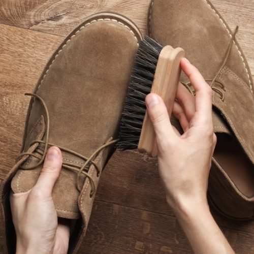 A person brushes a pair of brown leather shoes on a wooden floor as part of a simple at-home care routine.