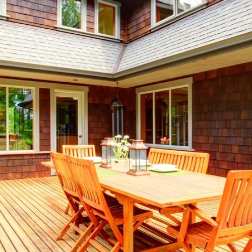 A wooden deck connected to the corner of a house. There is a large dining space in the middle, but the rest is quite open.