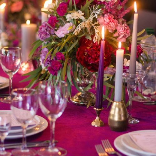 An elegant dining table with a hot pink tablecloth. Flowers, candles, and dinnerware line the table for an occasion.