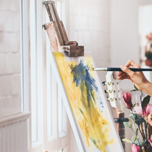 A woman creates an abstract blue and yellow painting on a canvas. She sits in a clean white room with natural lighting.