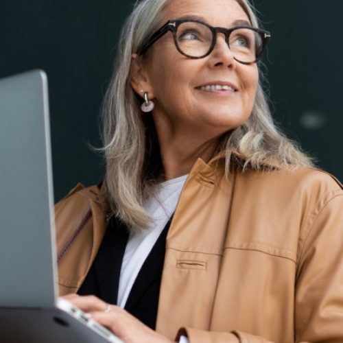 A woman with light gray hair standing in an office space and holding a laptop while smiling into the distance.