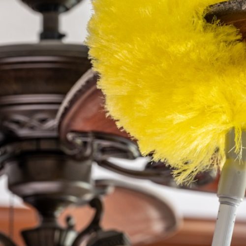 A close-up of a yellow duster wrapped around a ceiling fan blade. The fan blade is dusty and being cleaned.