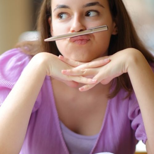 A young woman sits at her desk in a purple blouse with a pencil resting on her upper lip as she glares at the windows.