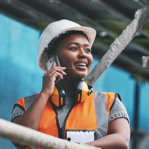 A woman in an orange safety vest and white hard hat stands on scaffolding and talks on a cell phone.