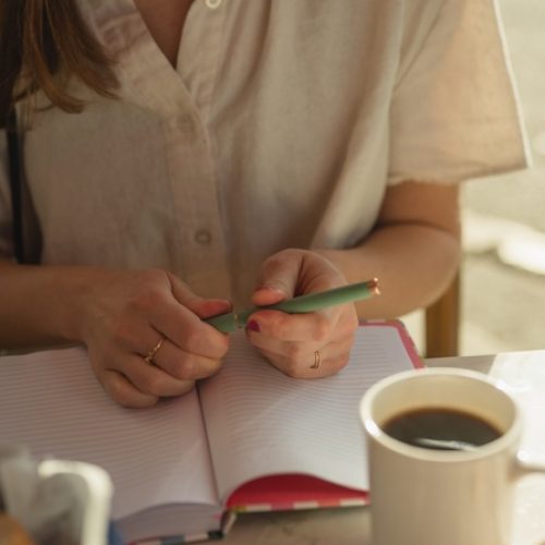 A close-up of someone sitting at a table with an open notebook in front of them. There is also a cup of black coffee.