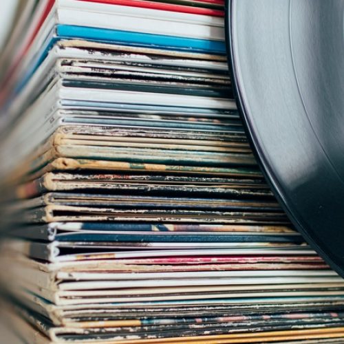A stack of vinyl records in their original album jackets sit in a pile on a white surface. An exposed vinyl leans against it.
