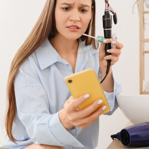 A woman checks her phone while curling her hair next to a laptop and hair dryer on a bathroom counter.
