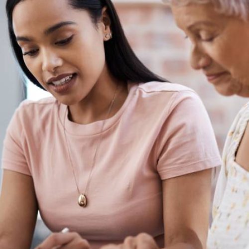 A woman reviews paperwork at a table with an older woman seated beside her as they look over the documents together.