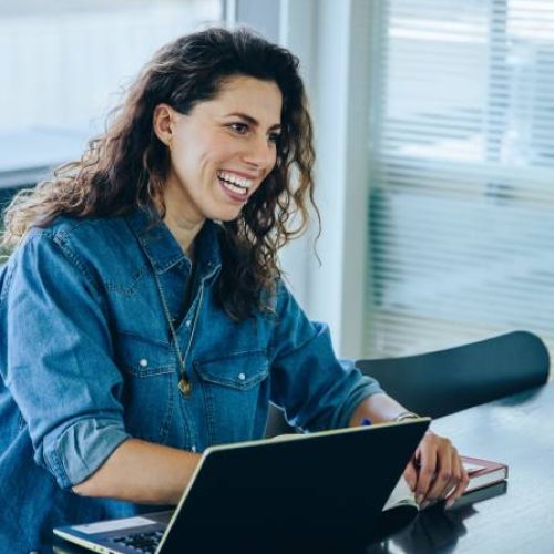 A smiling woman in business casual clothing sitting across a work table from a coworker. She has a laptop in front of her.