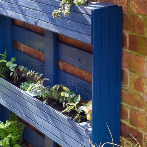 A vertical herb garden made out of a blue painted wooden pallet is leaning against a brick wall outdoors.