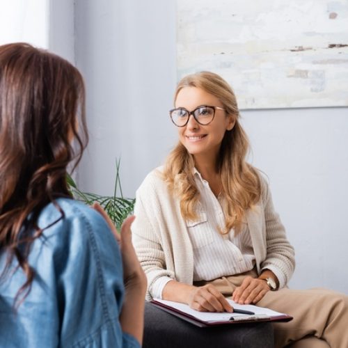A woman seen from behind sitting on a couch across from another woman holding a clipboard during a conversation.
