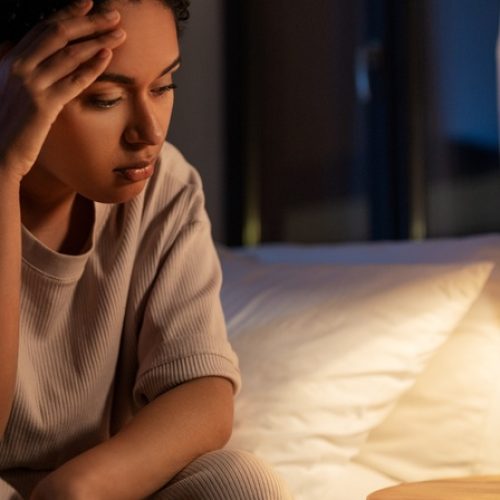 A woman sitting in her bed at night, holding her head with one hand while looking at the clock next to her table lamp.
