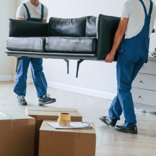 Two men wear denim overalls and white T-shirts as they carry a couch out of a home. There are boxes stacked up.