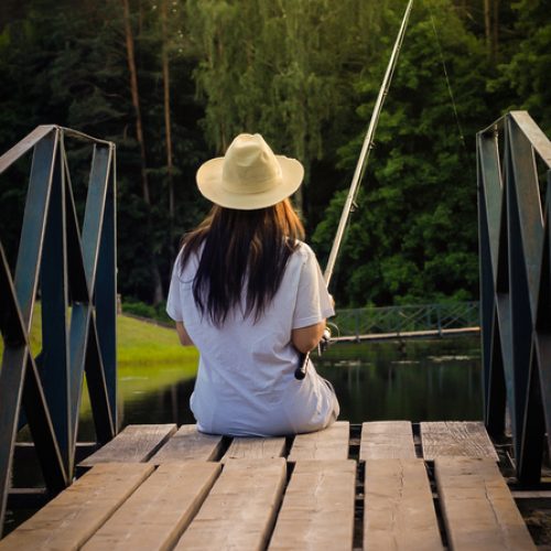 A woman in a white t-shirt and a light hat sits on the edge of a dock, fishing on a calm lake or pond with her hair down.