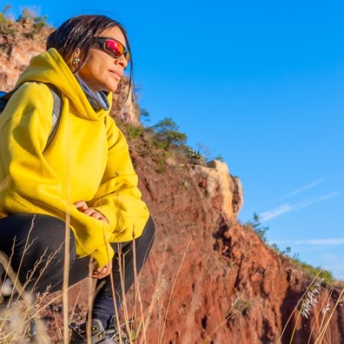 A hiker is wearing a yellow hoodie and backpack while squatting on a rocky hillside, overlooking a mountain landscape.