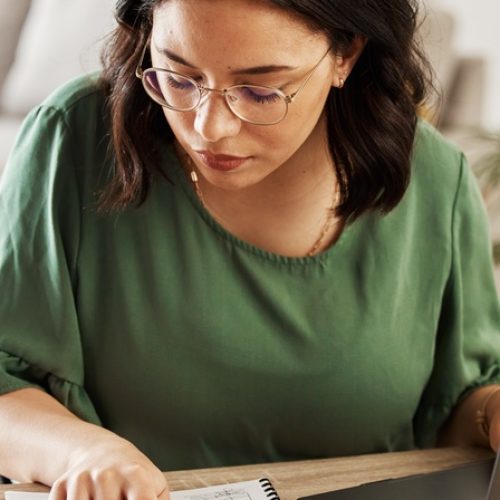 A woman sits at a desk in her home, comparing the financial information in her notebook to that on her computer.