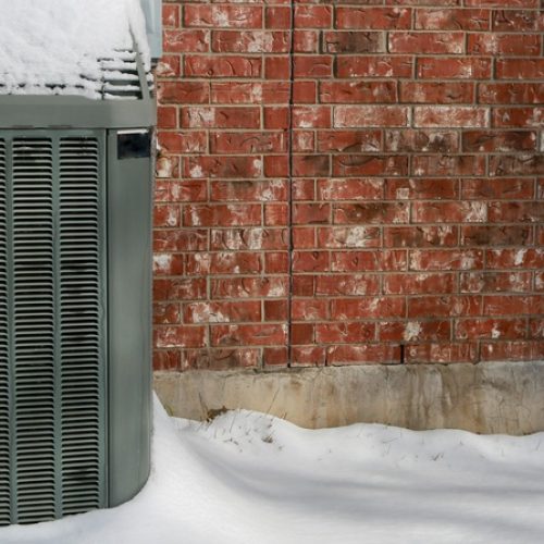 An outdoor AC unit sits against a brick house with snow on the ground surrounding it. There's also snow on the unit.