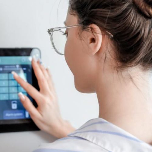 A woman using a home security screen that's attached to her wall. It has a camera graphic and a number pin pad.