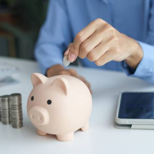 A person wearing a blue blouse is dropping a silver coin into the top opening of a pink piggy bank.