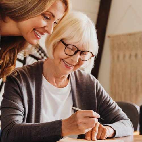 An older woman sitting down at the table to sign documents with her adult daughter over her shoulder to review them.