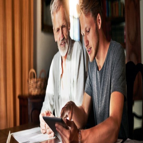 A father and son sitting at a table look at a tablet and discuss finances. The sun shines through a window behind them.