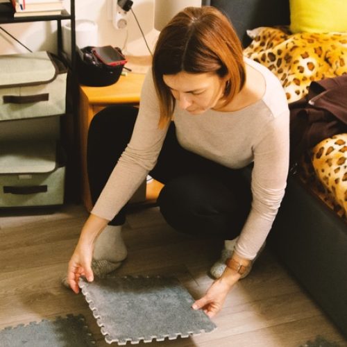 a woman crouching on a bedroom floor while assembling gray interlocking foam mats near a bed and storage shelves.